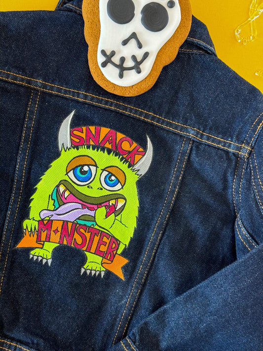 Stoofy hand-painted denim jacket for children featuring a green cartoon monster with a snack and text that reads 'SNACK MONSTER'. The jacket is blue denim with the monster design on the back. Design and copyright owned by UK artist, Stoofy