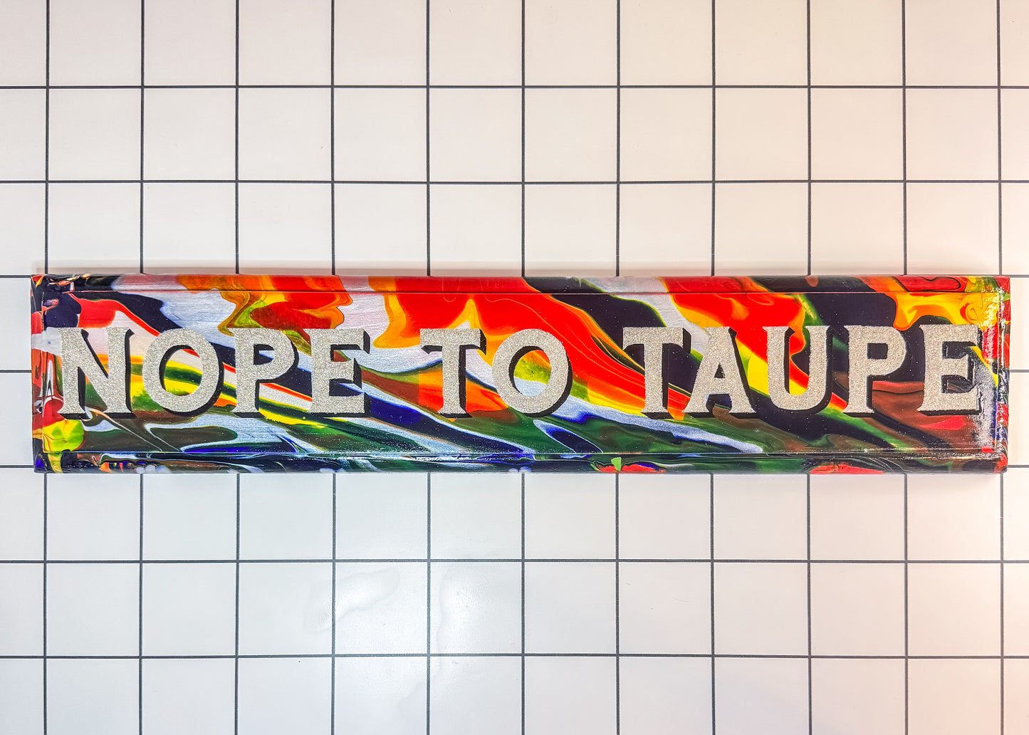 A hand-painted wooden sign with the phrase ‘NOPE TO TAUPE’ in bold, capital letters. The background features a swirling mix of vibrant colours, including red, yellow, green, blue, and black, creating a marbled effect. The text is painted in a contrasting light colour with a shadow effect, making it stand out against the chaotic, colourful backdrop. The sign is placed on a white tiled surface with black grid lines.