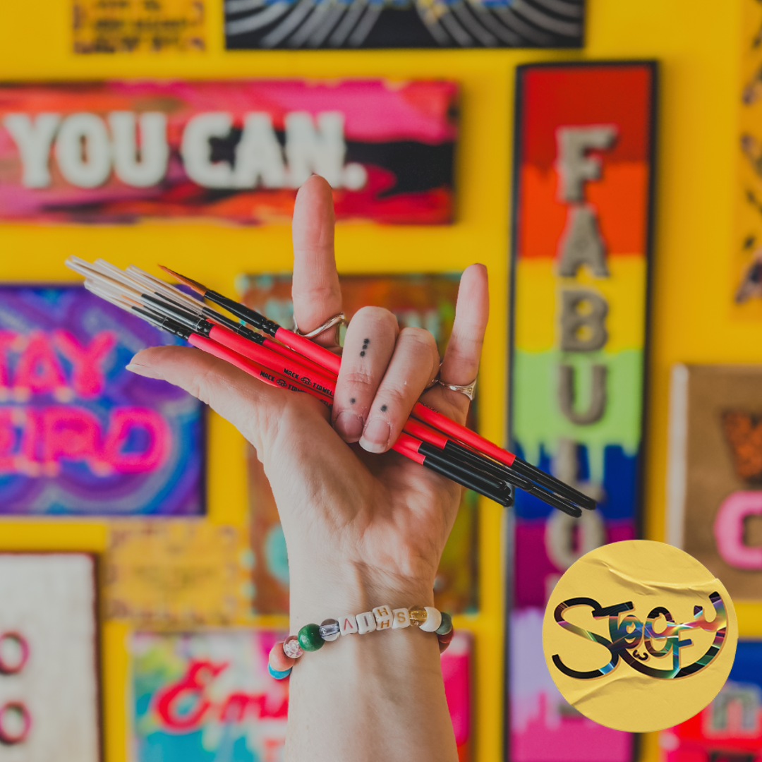 Stoofy UK artist holding paintbrushes in air in a rock and roll hand gesture in front of Stoofy's hand-painted sign collection display with Stoofy logo.
