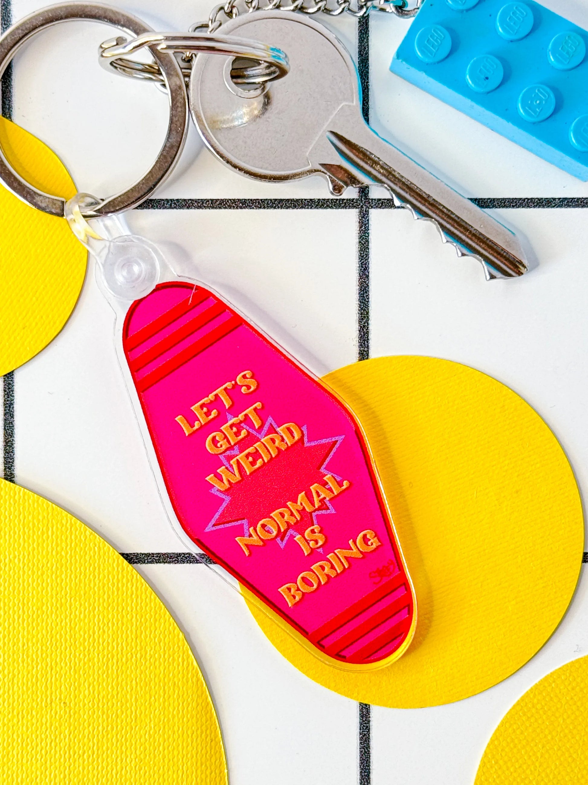 Stoofy's hot pink keyring with the text 'LET'S GET WEIRD' printed on it, displayed on a tiled surface with keys and a blue toy block in the background. Design and copyright owned by Stoofy.
