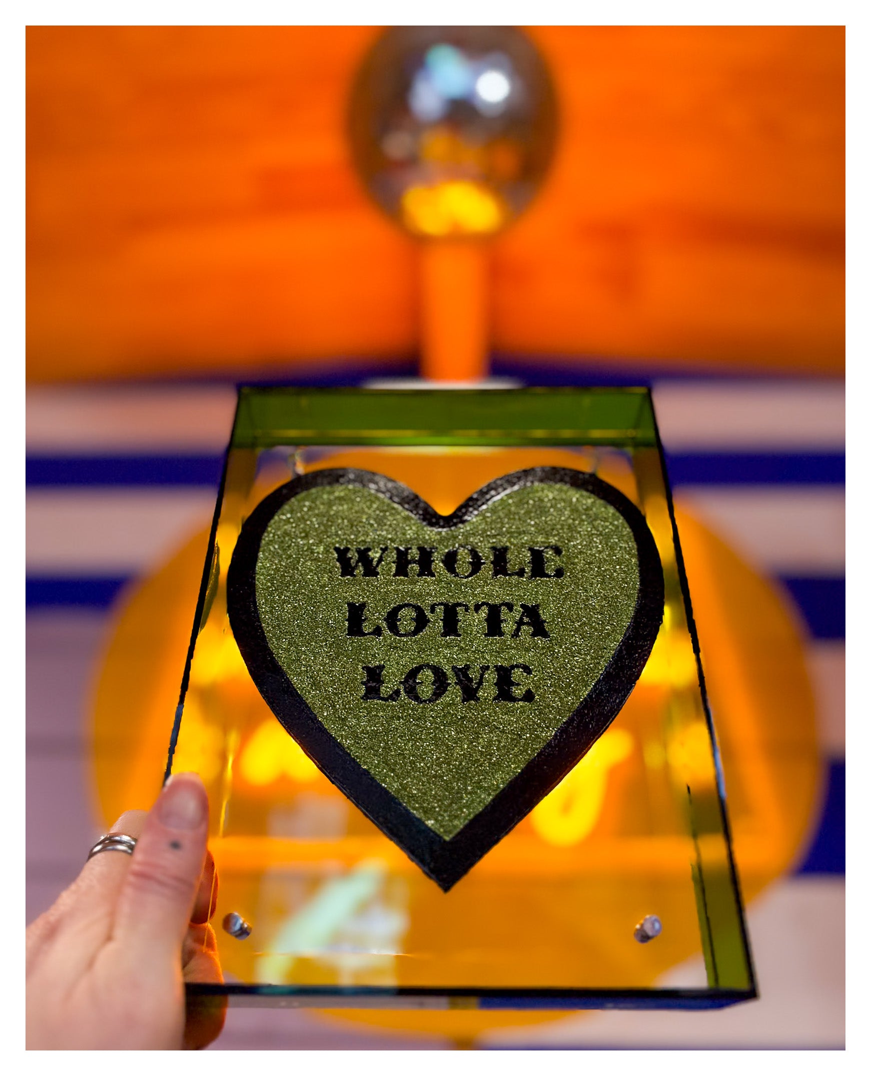 An artwork from Love is the Word Art Collection by contemporary UK Artist Stoofy. Heart-shaped hand-painted typography sign with 'Whole Lotta Love' text held by a hand against a colourful background.