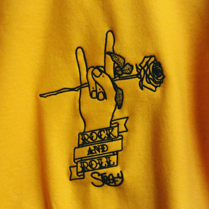 Close up product view of embroidered Stoofy design It’s Only R&R Baby! Unisex Sweatshirt in quality, heavyweight cotton, in gold with black embroidered design. Rock and roll hand gesture and rose. Perfect apparel choice for music lovers. Designed by UK, Artist Stoofy